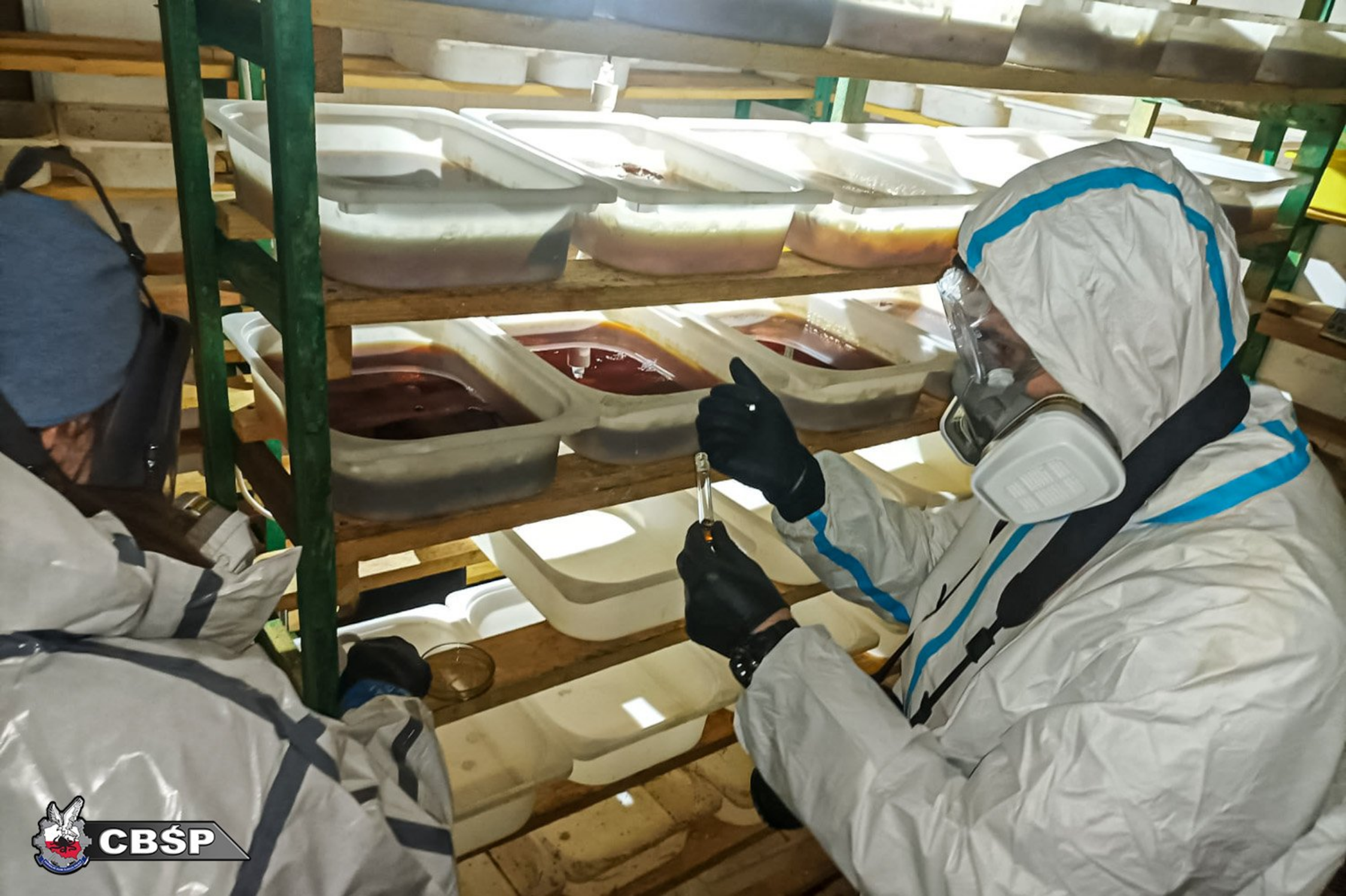 People in protective suits work with trays of liquid samples on metal shelves in a laboratory-like setting